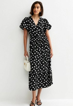 Kobieta ubrana w czarną sukienkę midi w białe kropki z bufkami na rękawach, trzymająca małą białą torebkę i nosząca czarne sandały na cienkich paskach.