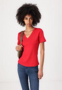 Tricou roșu cu mânecă scurtă și decolteu în V, realizat dintr-un material moale, prezentând un mic logo în partea stângă sus. Asortat cu blugi albaștri și o geantă de umăr verde.