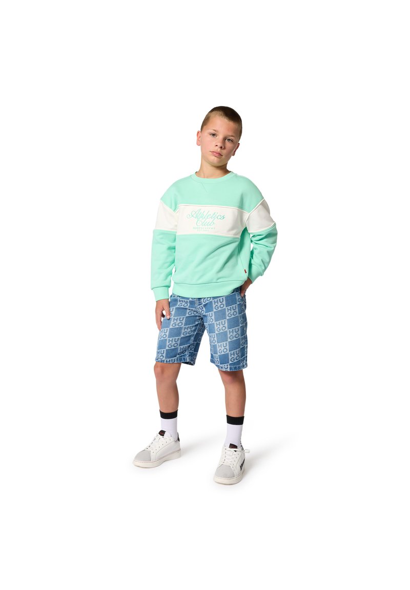 Ανοιχτό πρ άσινο φούτερ με λευκή ρίγα και λογότυπο, σε συνδυασμό με μπλε σορτς που έχουν επαναλαμβανόμενο μοτίβο λογότυπου. Λευκά αθλητικά παπούτσια και μαύρες κάλτσες.