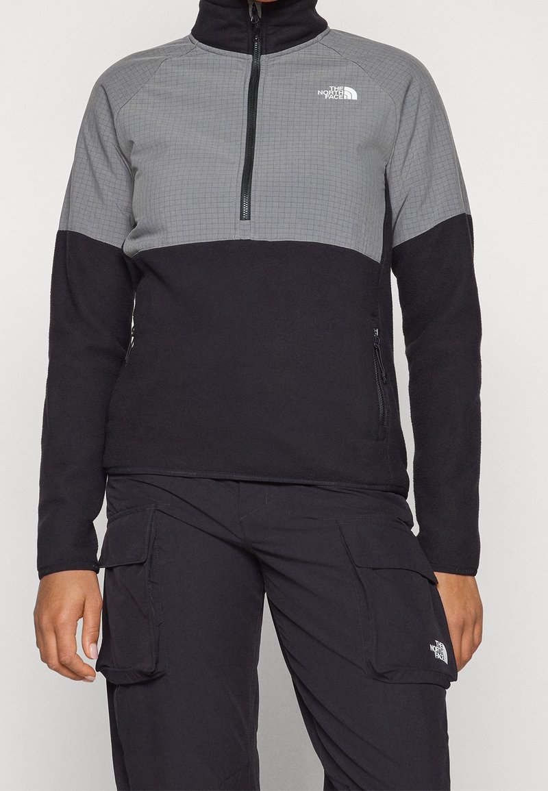 Persoon draagt een zwart-grijs fleecevest met rits en zwarte cargobroek met zijzakken, beide met het The North Face-logo.