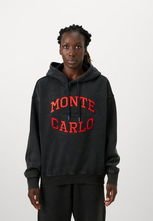 Brahuzott hajú nő, fekete kapucnis pulóverben és nadrágban, a pulóveren nagy vörös betűkkel ez áll: "MONTE CARLO".