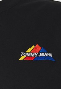 Svart t-shirt i tyg med ett broderat logo i gula, röda och blå trianglar med texten "TOMMY JEANS" i vitt nedanför.