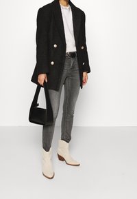 Blazer negro de doble botonadura con botones dorados, suéter ligero, jeans grises, botas de vaquero blancas y un bolso negro. Texturas suaves y ajuste a medida.