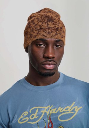 Hombre de piel oscura lleva un gorro de lana marrón con estampado y una camiseta azul Ed Hardy con texto amarillo y un gráfico colorido debajo del texto.