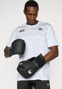 Μαύρα γάντια πυγμαχίας με ιμάντες βέλκρο, φορεμένα από άτομο που φοράει λευκό αθλητικό πουκάμισο με μοτίβο. Το πουκάμισο έχει γεωμετρικό σχέδιο και λογότυπο.