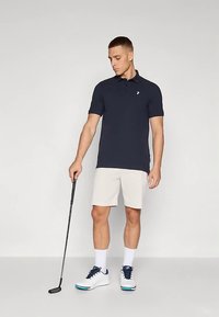 Mörkblå pikétröja med korta ärmar och en liten logotyp, kombinerad med ljusbeige shorts. Modellen håller i en svart golfklubba och bär vita skor.
