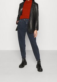 Blazer di pelle nera sopra un dolcevita rosso, abbinato a pantaloni in denim scuri e stivaletti neri con tacco. Stilizzato con una vestibilità comoda.