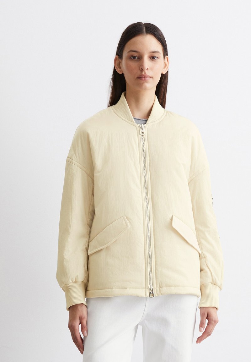 Veste bomber beige avec fermeture éclair à l'avant, poignets côtelés et poches latérales. Présente une texture douce et lisse ainsi qu'une coupe décontractée.