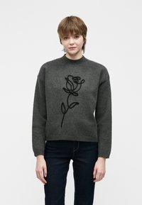 Jovem com cabelo curto castanho, vestindo um suéter cinza escuro com um design abstrato de rosa preta, combinado com jeans escuros, de pé contra um fundo branco.