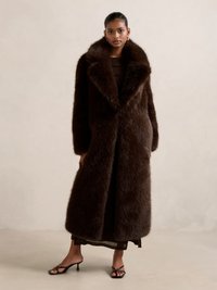 Brun faux fur kåpe med bred krage, lange ermer og en fyldig silhuett. Båret over en mørk kjole med en glatt tekstur.