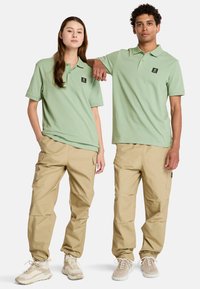 Gröna bomullspikéer med kort ärm och bröstlogotyp, kombinerat med beigefärgade cargobyxor och ljusa sneakers.