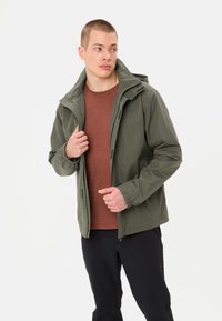 Waterdichte groene jas met capuchon, rits aan de voorkant, elastische manchetten en verstelbare zoom, gecombineerd met een bruine shirt en zwarte broek.