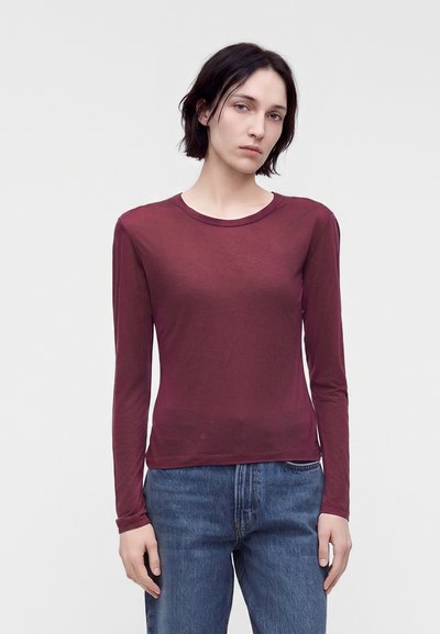 Femme aux cheveux courts et foncés portant une chemise bordeaux à manches longues et un jean bleu, debout devant un fond clair uni.