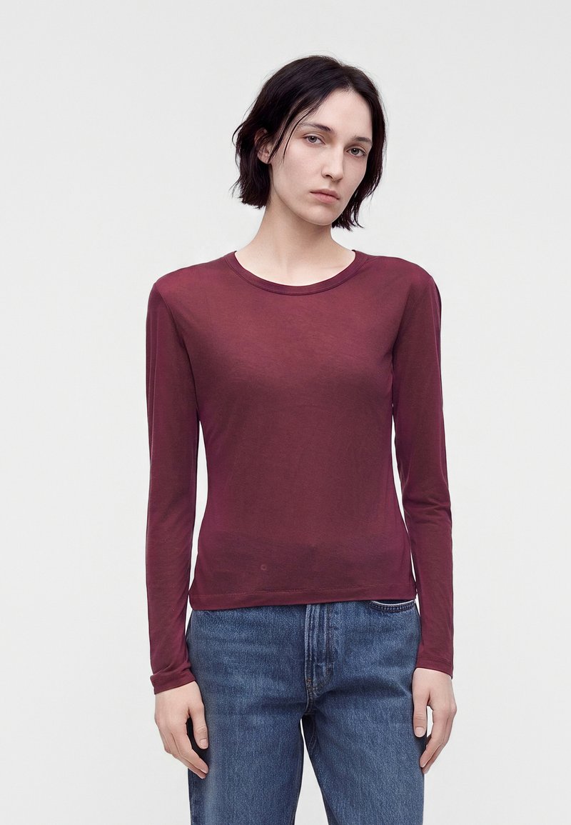 Femme aux cheveux courts et foncés portant une chemise bordeaux à manches longues et un jean bleu, debout devant un fond clair uni.