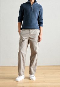 Maglione blu navy a coste con collo a mezza zip, abbinato a pantaloni larghi beige chiaro e sneakers bianche su un pavimento di legno.