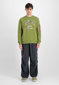 Felpa verde oliva con grande logo "ALPHA", colletto a giro, polsini a coste e tasca con zip sulla manica, abbinata a pantaloni scuri e larghi.