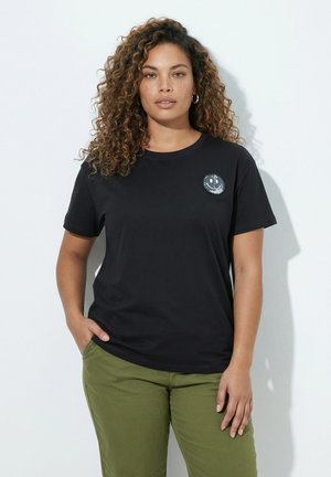 Woman with curly hair wearing a black t-shirt with a smiley face patch and olive green pants, standing with one hand in pocket.