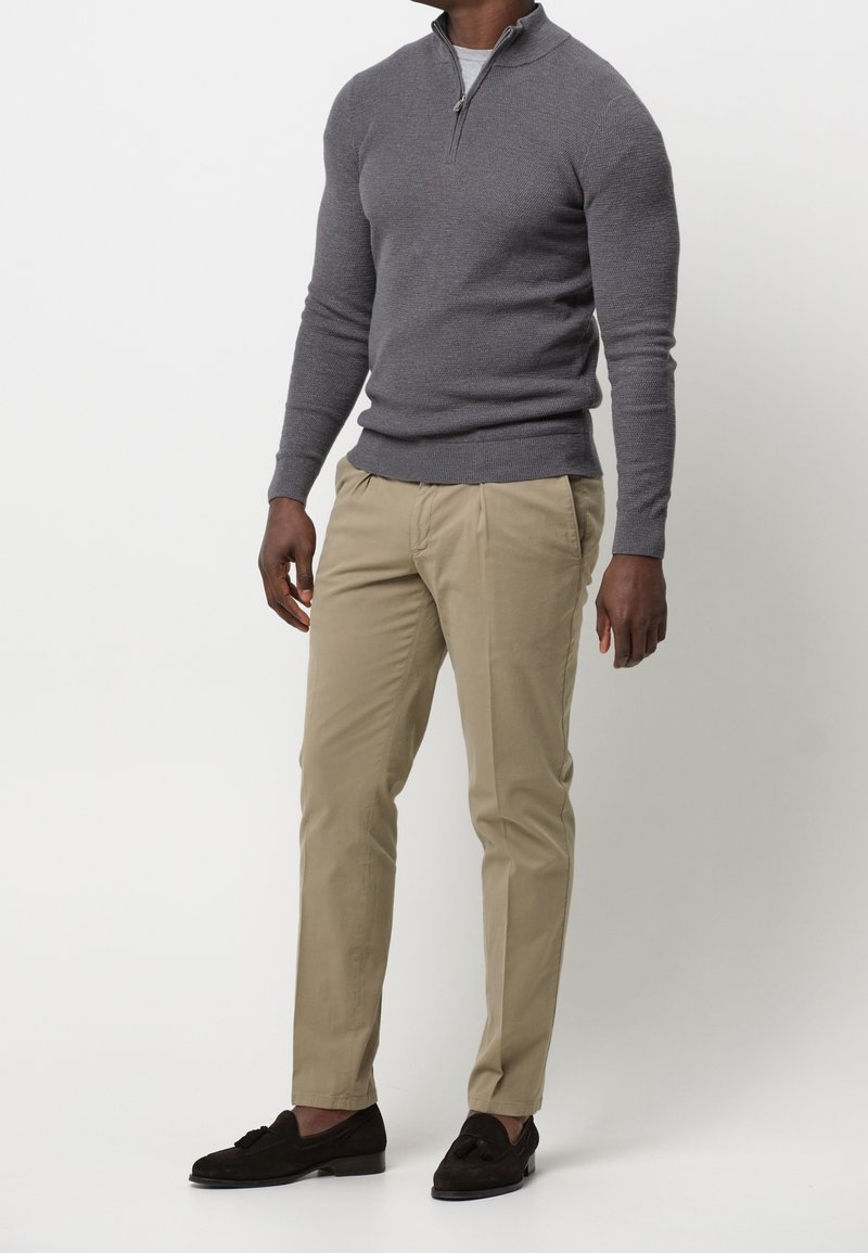 Homme portant un pull gris à demi-fermeture éclair, un pantalon chino beige et des mocassins marron foncé à glands, debout devant un fond blanc uni.