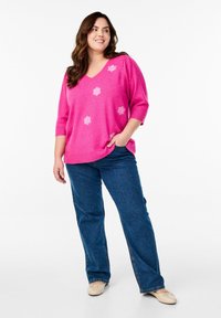 Rosa Strickpullover mit V-Ausschnitt und kurzen Ärmeln, verziert mit weißen Blumenstickereien. Kombiniert mit blauen, geraden Jeans und cremefarbenen Ballerinas.