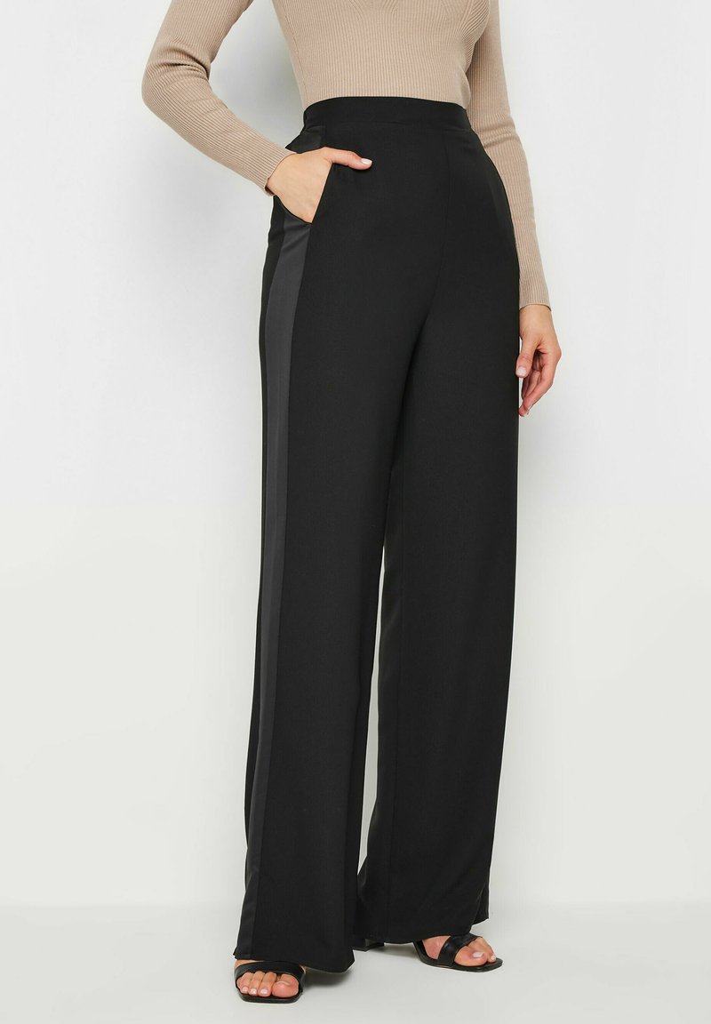 Pantalons larges noirs avec une bande de satin sur le côté, présentant une taille haute et des poches latérales. Tissu lisse avec une coupe structurée.