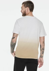 Man med kort brunt hår och tatueringar på båda armarna, iklädd en vit-beige gradient t-shirt och svarta byxor, vänd bort från kameran.