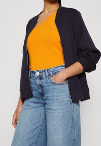 Woman wearing a bright orange sleeveless top, navy blue zip-up jacket with sleeves pushed up, and light blue high-waisted jeans.