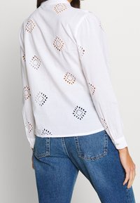 Blouse blanche à manches longues avec motifs découpés brodés en forme de losange, associée à un jean en denim bleu.