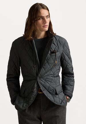 Man with shoulder-length hair wearing a dark quilted jacket over a black sweater, with sunglasses in the chest pocket and hands in pockets.