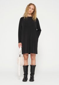 Vestido negro de manga larga con un corte holgado, que presenta bolsillos laterales. Combinado con botas negras hasta la rodilla y un bolso de piel sintética blanco.