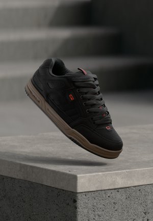 Baskets noires avec semelle marron, accents rouges et lacets épais suspendues au-dessus d'un rebord en béton avec des marches en arrière-plan.