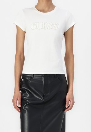 Valkoinen puuvillapaita lyhyillä hihoilla, jossa on aaltoileva tekstuurinen "GUESS"-logo. Yhdistetty mustaan nahkatehtailevaan hameeseen.