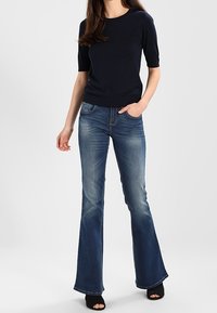 Suéter corto de color azul marino combinado con jeans acampanados de color azul. Los jeans tienen un aspecto desgastado y un dobladillo deshilachado; el modelo lleva unos zapatos de tacón negro abiertos por delante.