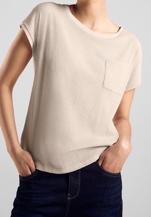 Beige top met korte mouwen en een textuureffect, voorzien van een borstzak, gecombineerd met donkere spijkerbroek. Afgeronde halslijn en een losse pasvorm.