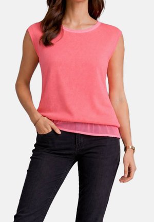 Femme portant un haut sans manches rose avec un détail en tulle à l'ourlet et un jean noir, main dans la poche, sur un fond uni.