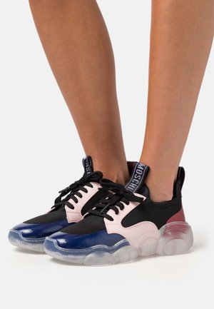 Piernas con zapatillas negras, rosas y azules con suelas gruesas y transparentes y cordones negros, de pie sobre una superficie blanca con un fondo liso.