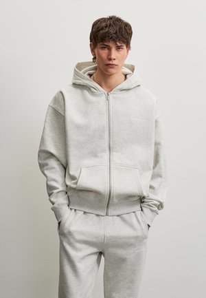 Jeune homme aux cheveux courts bruns portant un sweat à capuche gris clair zippé et un pantalon de survêtement assorti, debout les mains dans les poches devant un fond uni.