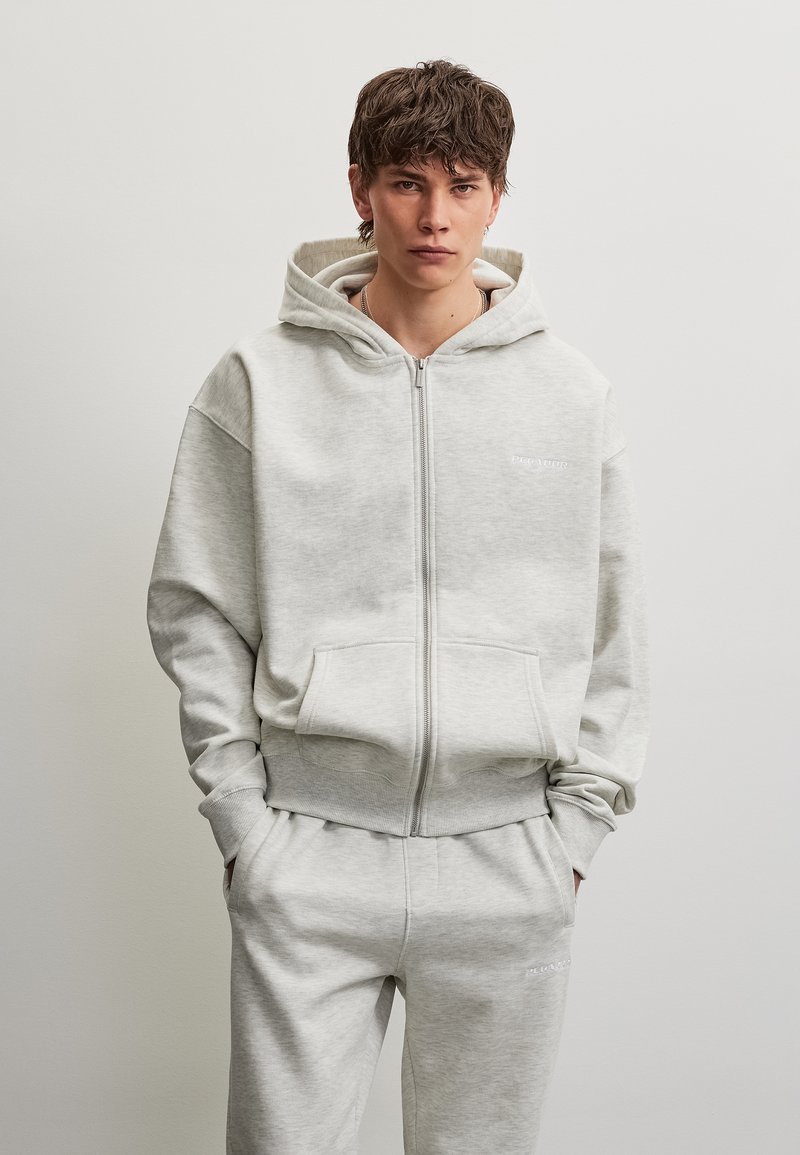 Jeune homme aux cheveux courts bruns portant un sweat à capuche gris clair zippé et un pantalon de survêtement assorti, debout les mains dans les poches devant un fond uni.