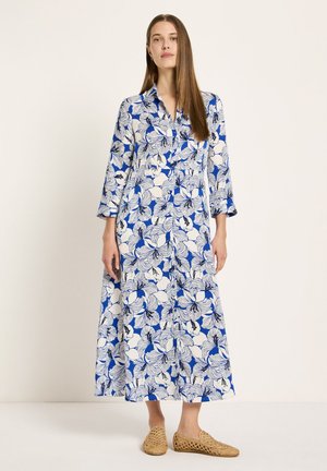 Frau in einem langen blau-weißen Blumenkleid mit dreiviertel Ärmeln und beigen gewebten Schlupfschuhen, stehend vor einem schlichten Hintergrund.