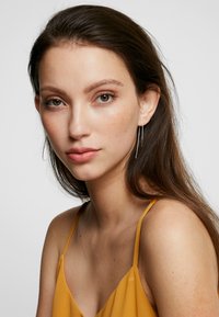Camisole en satin doré avec des fines bretelles, présentant une texture lisse. Le mannequin a de longs cheveux foncés et porte des boucles d'oreilles argentées longues.