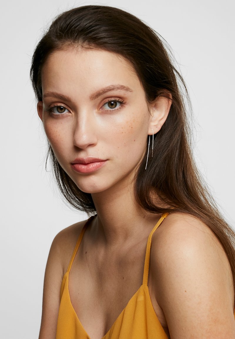 Camisole en satin doré avec des fines bretelles, présentant une texture lisse. Le mannequin a de longs cheveux foncés et porte des boucles d'oreilles argentées longues.