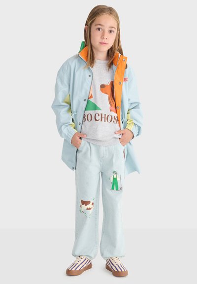 Enfant portant une veste bleu clair avec une doublure orange, un sweat-shirt gris avec un motif de chien, un pantalon bleu clair orné de patchs fermier et vache, chaussures rayées.