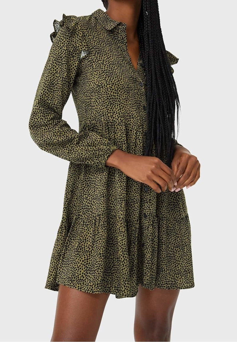 Vestido verde oliva con patrón de manchas negras, mangas largas, cuello de volantes y cierre frontal con botones. La falda acampanada y en capas aporta volumen.
