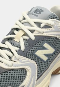 Gray mesh running shoe featuring a padded tongue, cream laces, and a prominent light gray "N" logo on the side. Textured rubber sole.