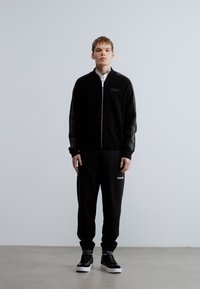 Casaco preto com fecho éclair e logótipo, combinado com calças de treino pretas com um pequeno logótipo. Modelo usa ténis pretos. Textura lisa em toda a peça.