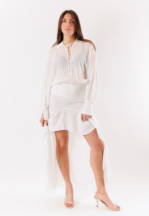 Donna che indossa una camicetta bianca trasparente a maniche lunghe e una gonna bianca asimmetrica, in piedi con una gamba in avanti, sandali beige con tacco.