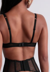 Bralette en dentelle noire avec bretelles ajustables et fermeture à crochet. Tissu transparent avec un motif géométrique et des détails à bord festonné.