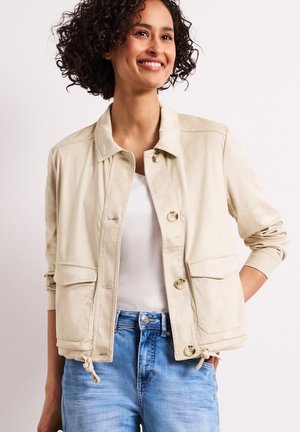 Uśmiechnięta kobieta z kręconymi włosami, ubrana w beżową kurtkę zapinaną na guziki na białą koszulę i niebieskie dżinsy, stojąca z jedną ręką w kieszeni.