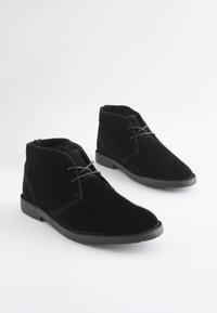 Paar schwarze Wildleder-Stiefeletten mit Schnürverschluss und Gummisohlen auf weißem Hintergrund.