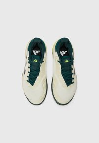 Gaiši krēmi treniņu apavi ar dziļi zaļiem akcentiem, ar fakturētu sieta augšdaļu, saites sistēmu un rakstainu gumijas zoli. Iespējama Adidas zīmola identificēšana.