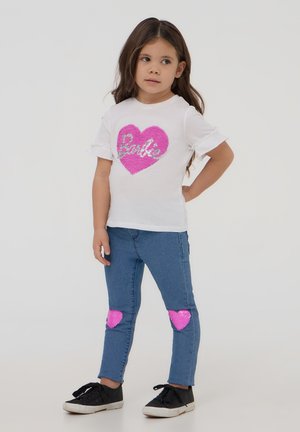 T-shirt bianca con cuore di paillettes rosa e testo "Barbie", abbinata a jeans di denim blu con toppe a cuore rosa sulle ginocchia e sneakers nere.